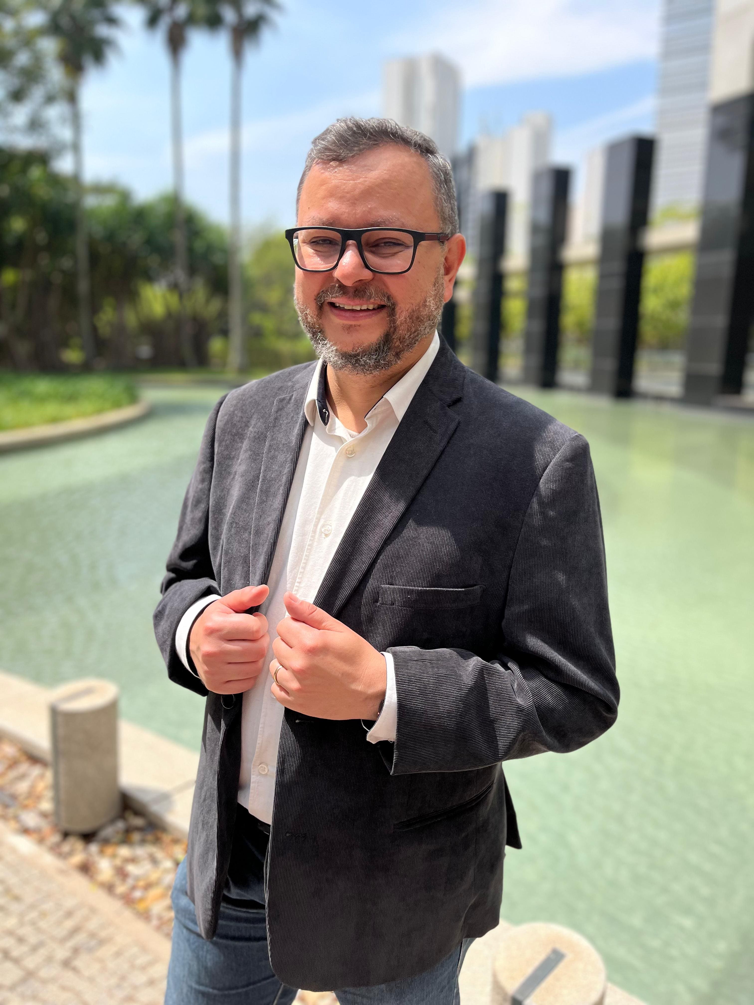 Homem branco com cabelos e barba grisalhos. Ele veste uma camisa social branca e um blaser cinza escuro. Ao fundo, parte de um grande edifício comercial rodeado de paisagismo e uma lagoa artificial.